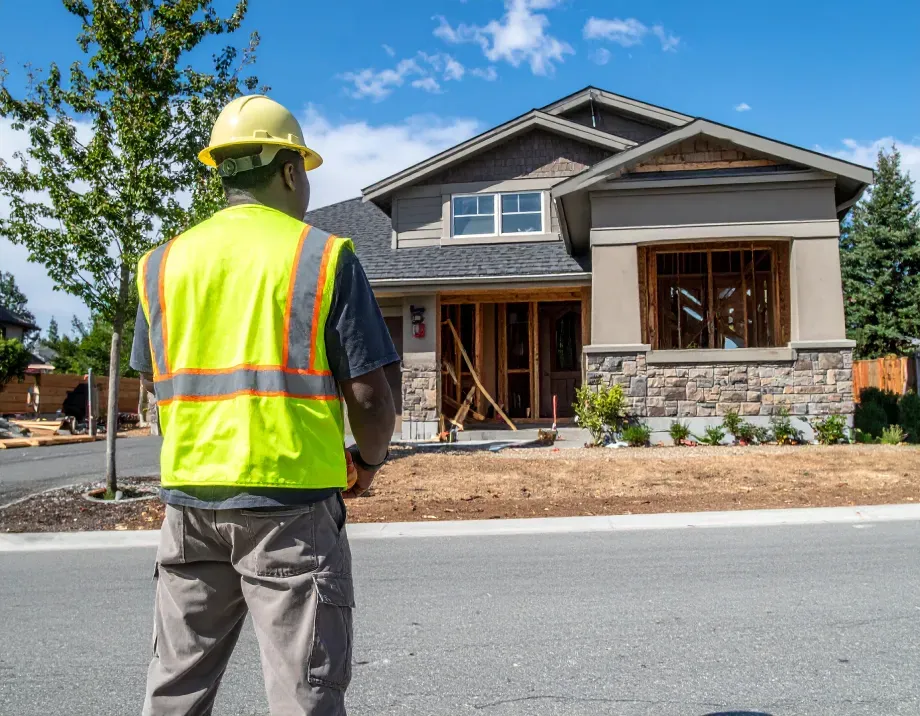 Construction worker in yellow vest and hard hat surveying a house under construction.
