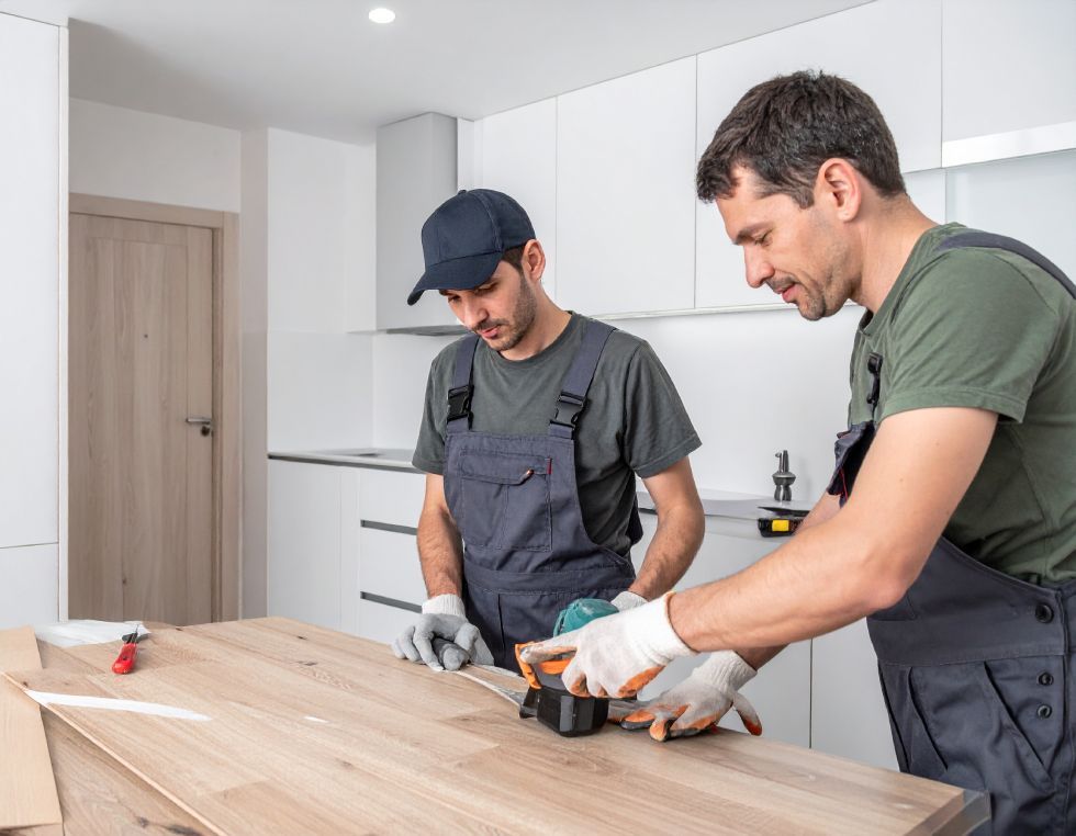 Two workers install wooden flooring in a white kitchen, one using a power sander.