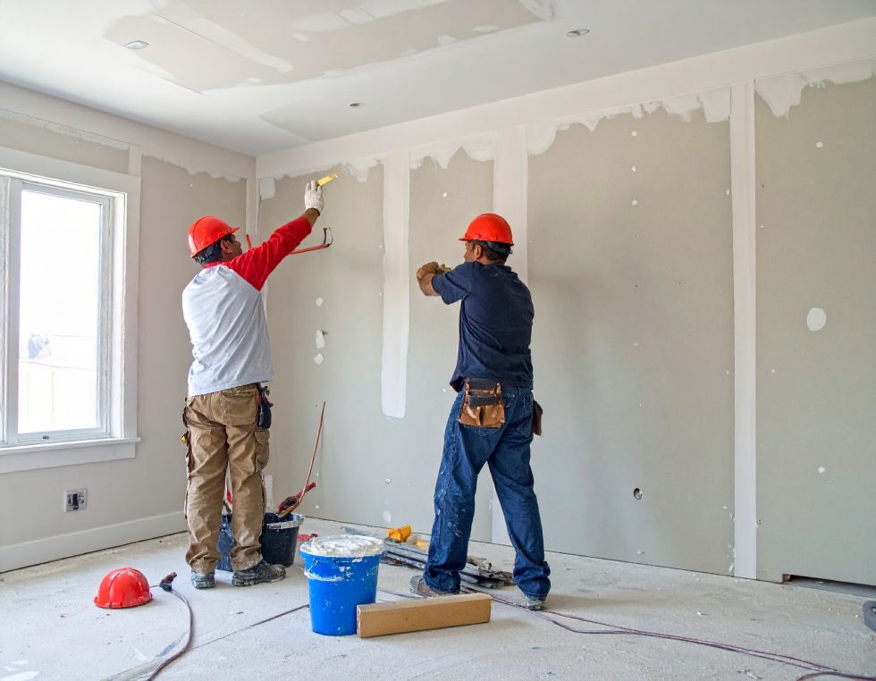 Two construction workers in hard hats working on drywall in a room.