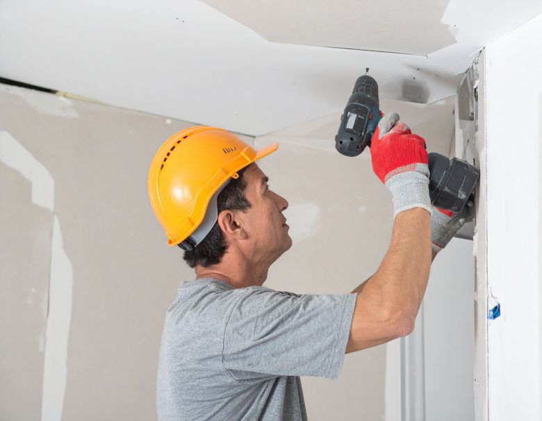 Construction worker drills into a wall while wearing a hard hat and gloves.