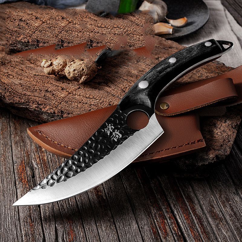 A hammer-textured boning knife with a black handle rests on a brown leather sheath atop a rustic wood surface.