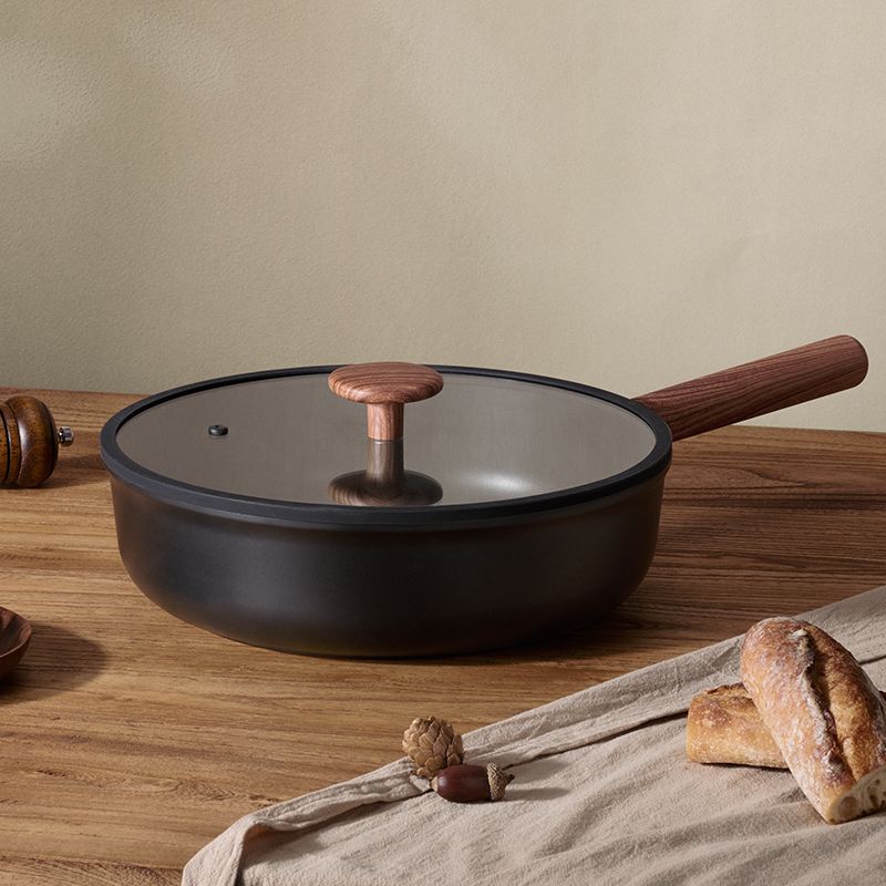 A black pan with a glass lid and wooden handle rests on a wooden table near a linen cloth and pieces of bread.