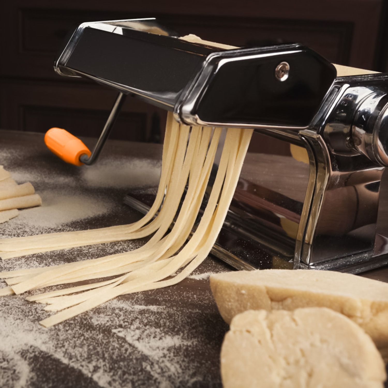 A stainless steel pasta machine with an orange-handled crank and table clamp, shown alongside samples of finished pasta.