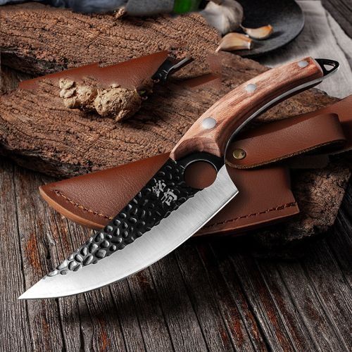 A textured, hammer-forged knife with a wooden handle rests on a brown leather sheath atop a weathered wooden surface.