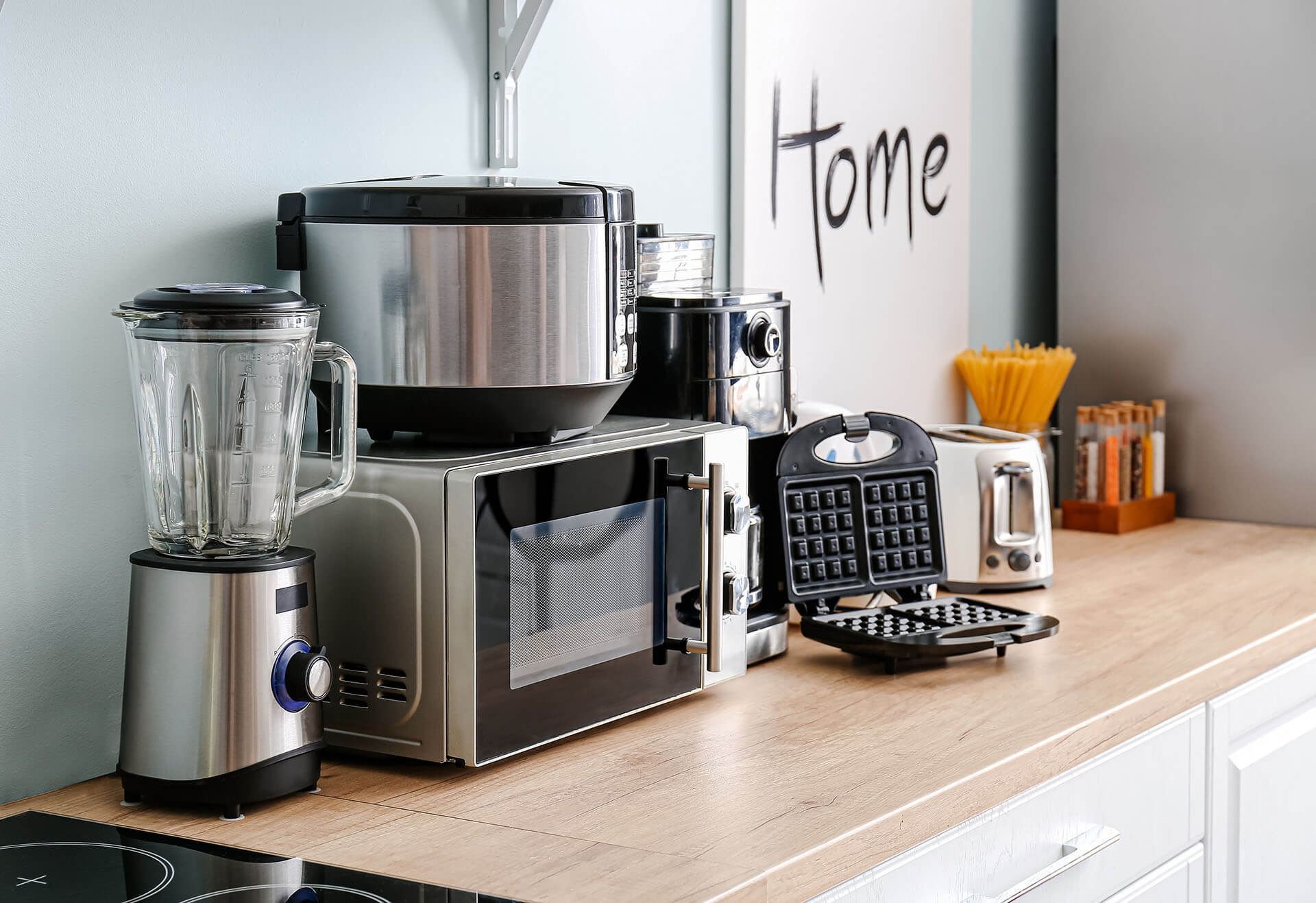 Kitchen countertop with various stainless steel appliances, including a blender, microwave, rice cooker, and waffle maker.