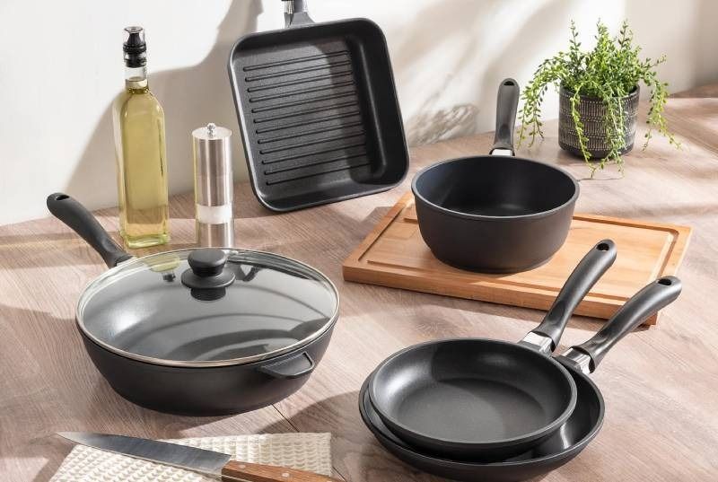 A collection of black non-stick cookware including pans, a griddle, and a pot arranged on a wooden table with accessories.