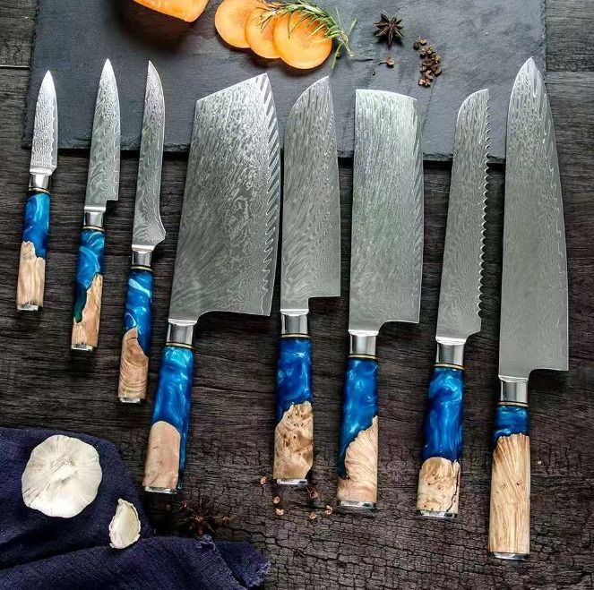 Eight kitchen knives with blue resin and wood handles and Damascus steel blades arranged on a dark wooden surface.