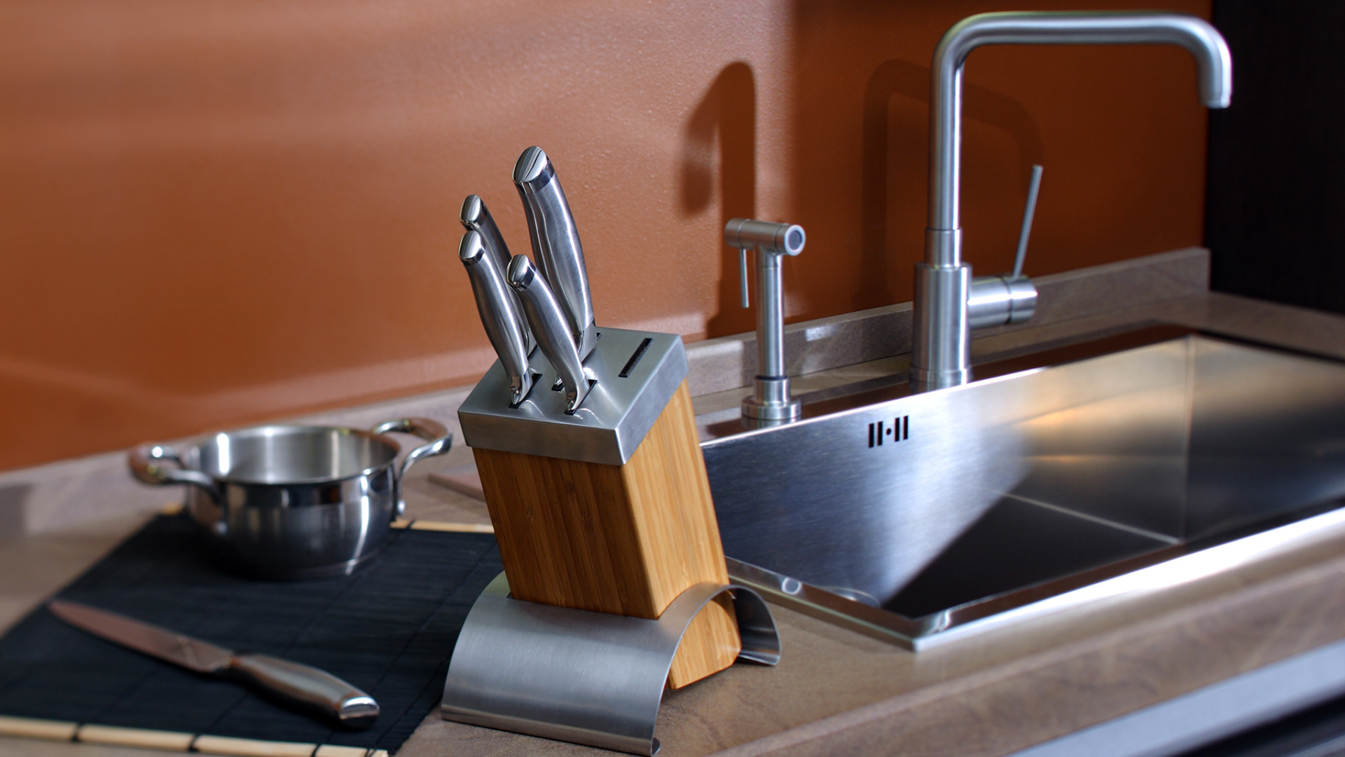A wooden knife block with several knives, a loose knife, and a small metal pot sit beside a kitchen sink and faucet.