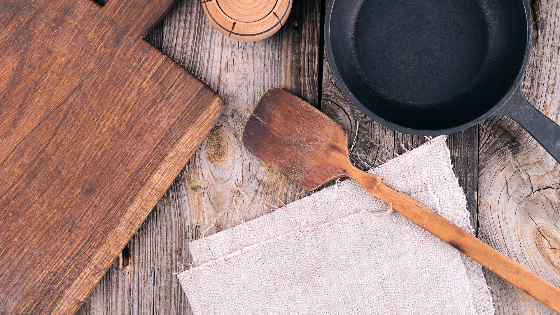 A cast-iron skillet, wooden spatula, and cutting board arranged on a rustic, weathered wooden table with a linen cloth.