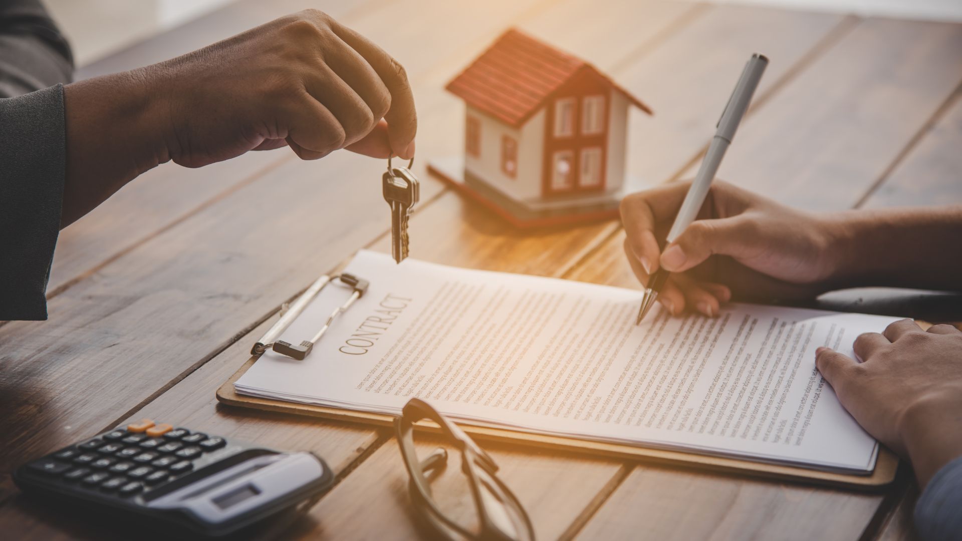 Hands signing a real estate document beside a calculator and small house model on a desk