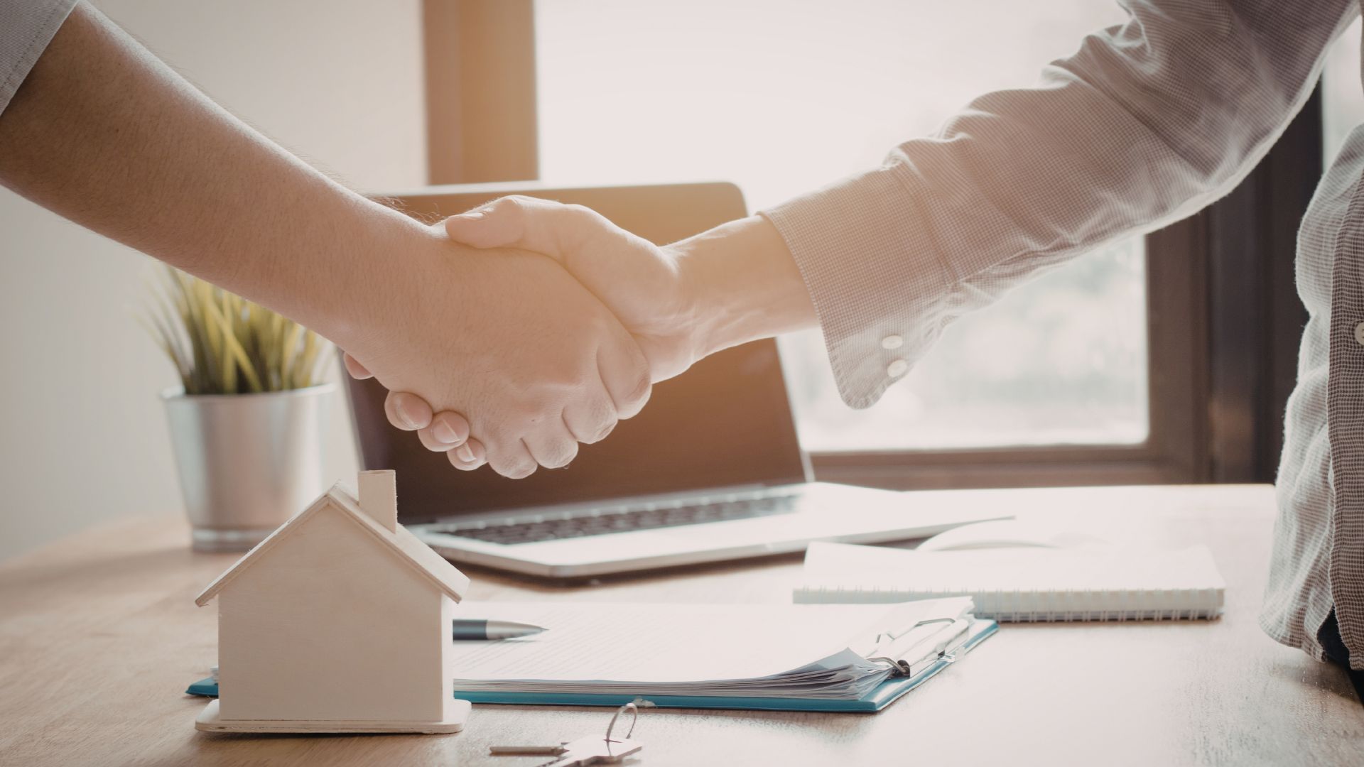 Two people shake hands across a desk with a small house model and laptop in a bright office