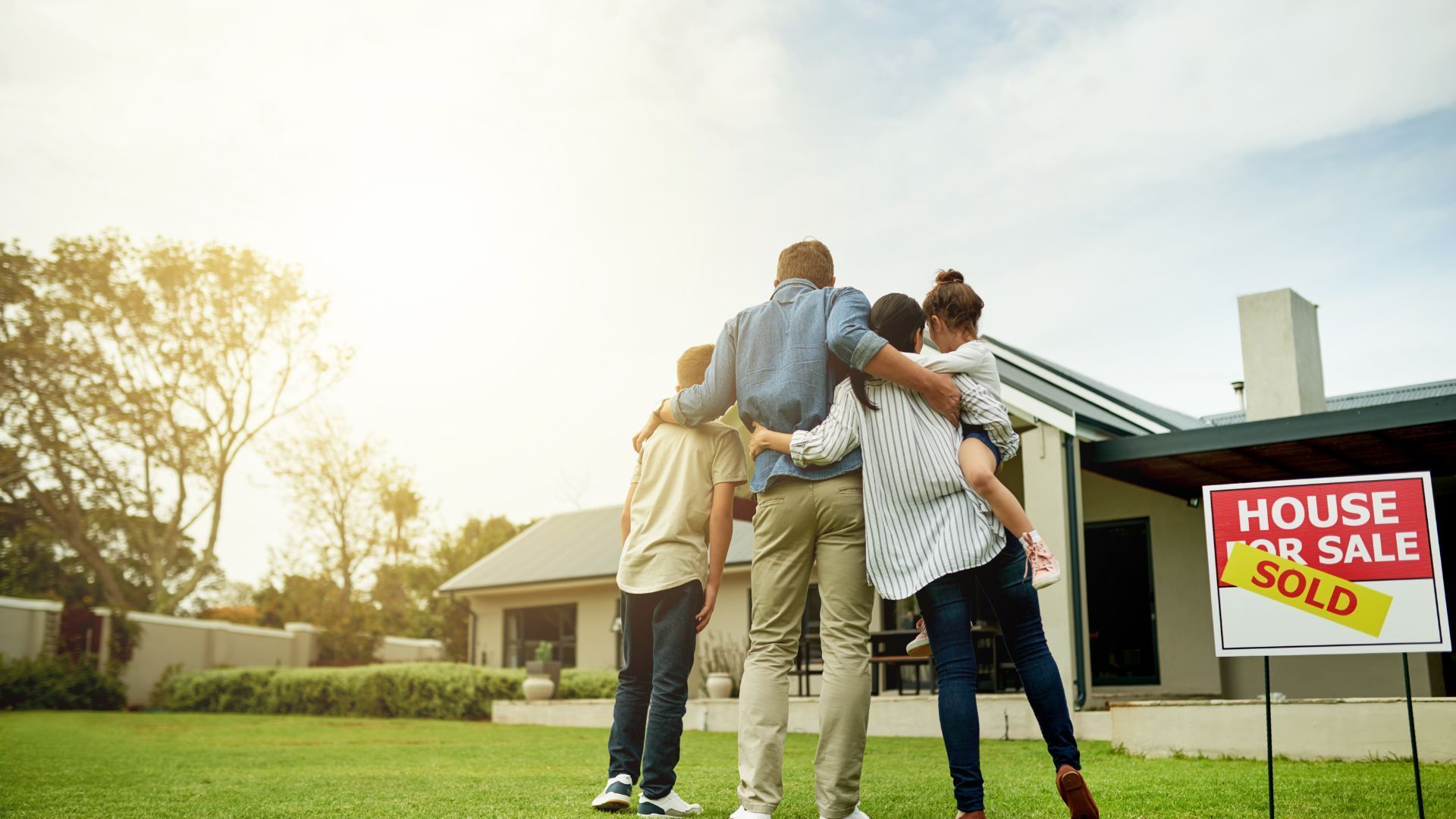 Family stands with arms around each other in front of a house with a “Sold” sign.