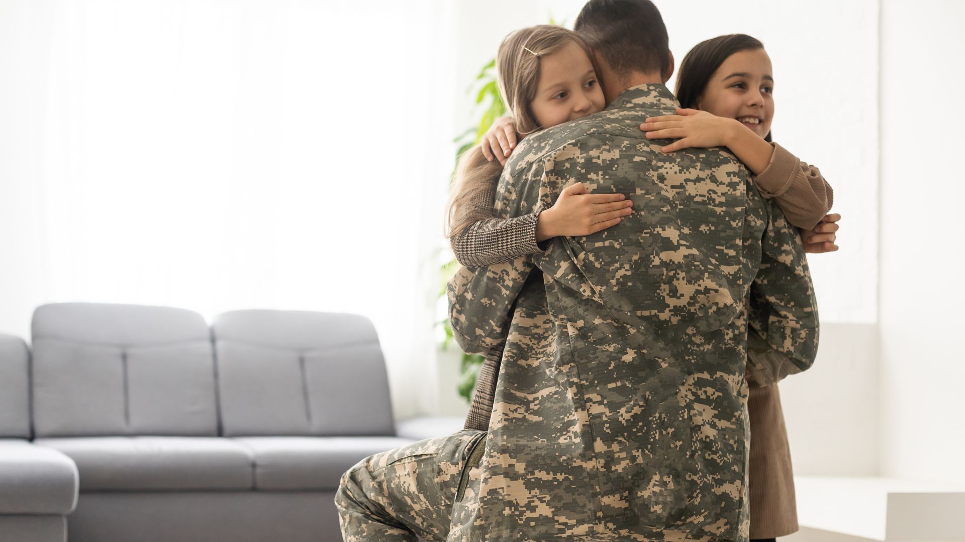 Service member embraces two smiling children in a bright living room with a gray sofa.