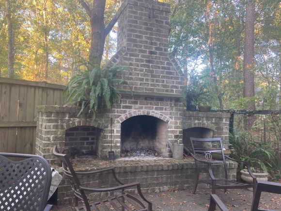 A brick fireplace is surrounded by chairs and trees in a backyard.