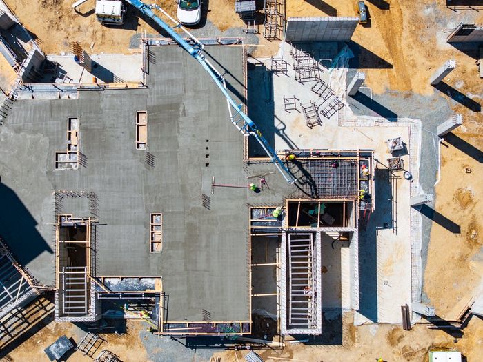 Aerial view of a construction site with a freshly poured concrete foundation, showing the building's footprint, workers, and a concrete pump.