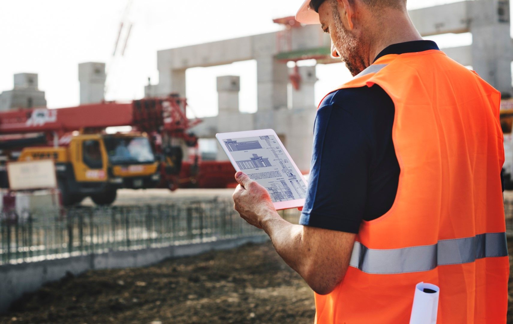 Construction worker in orange vest, helmet, reviewing plans on a tablet at a construction site with a crane and building in the background.