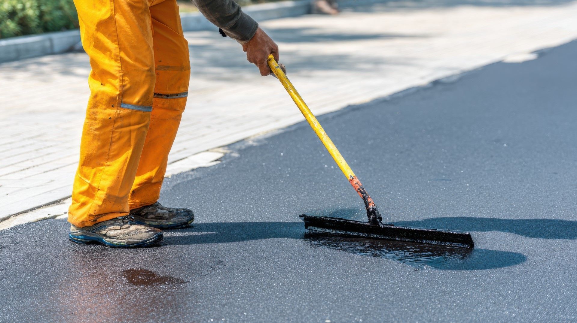 Person in orange work pants smoothing fresh black asphalt with a squeegee in daylight.