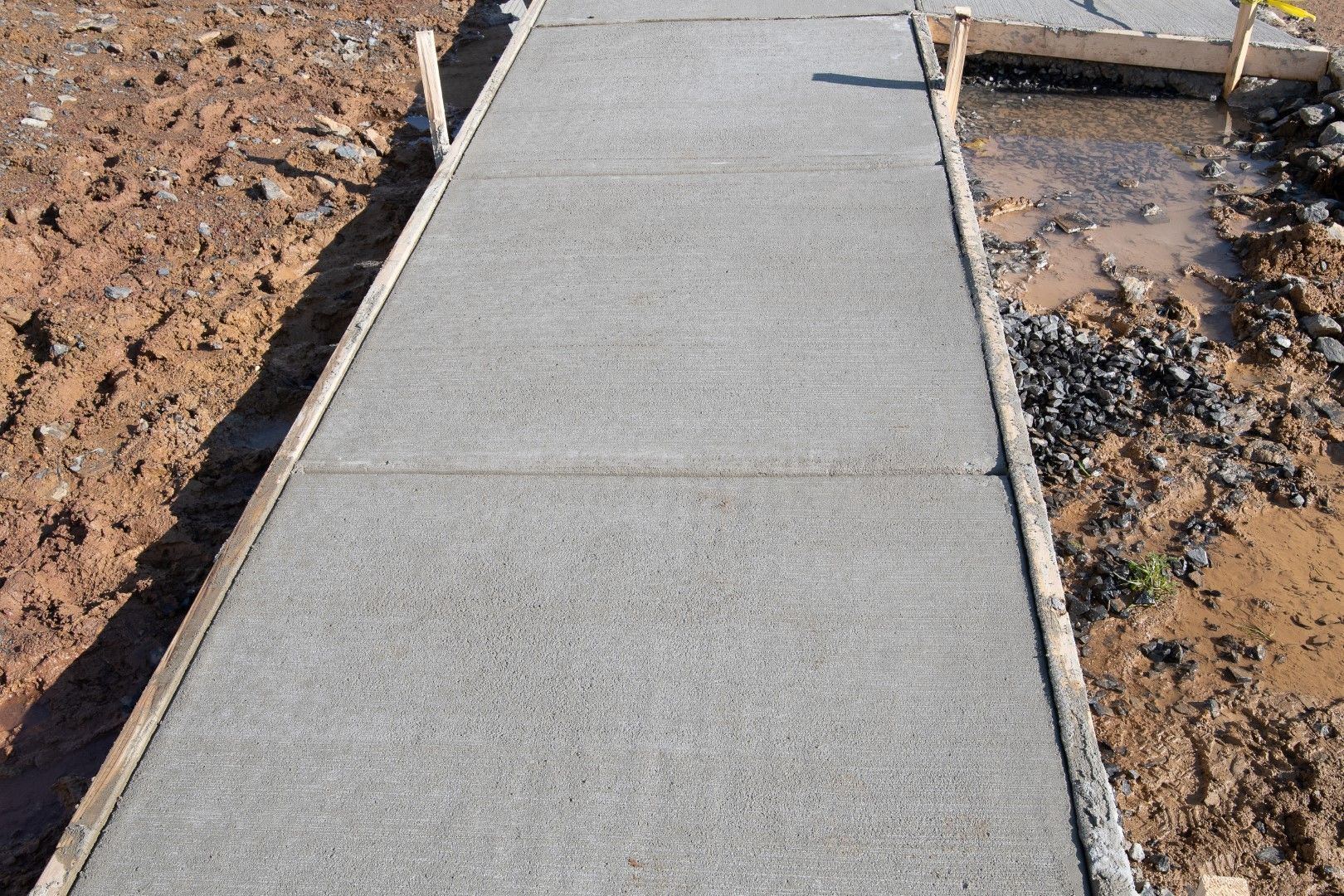 Freshly poured concrete sidewalk, framed by wooden forms, in a construction setting with surrounding dirt.
