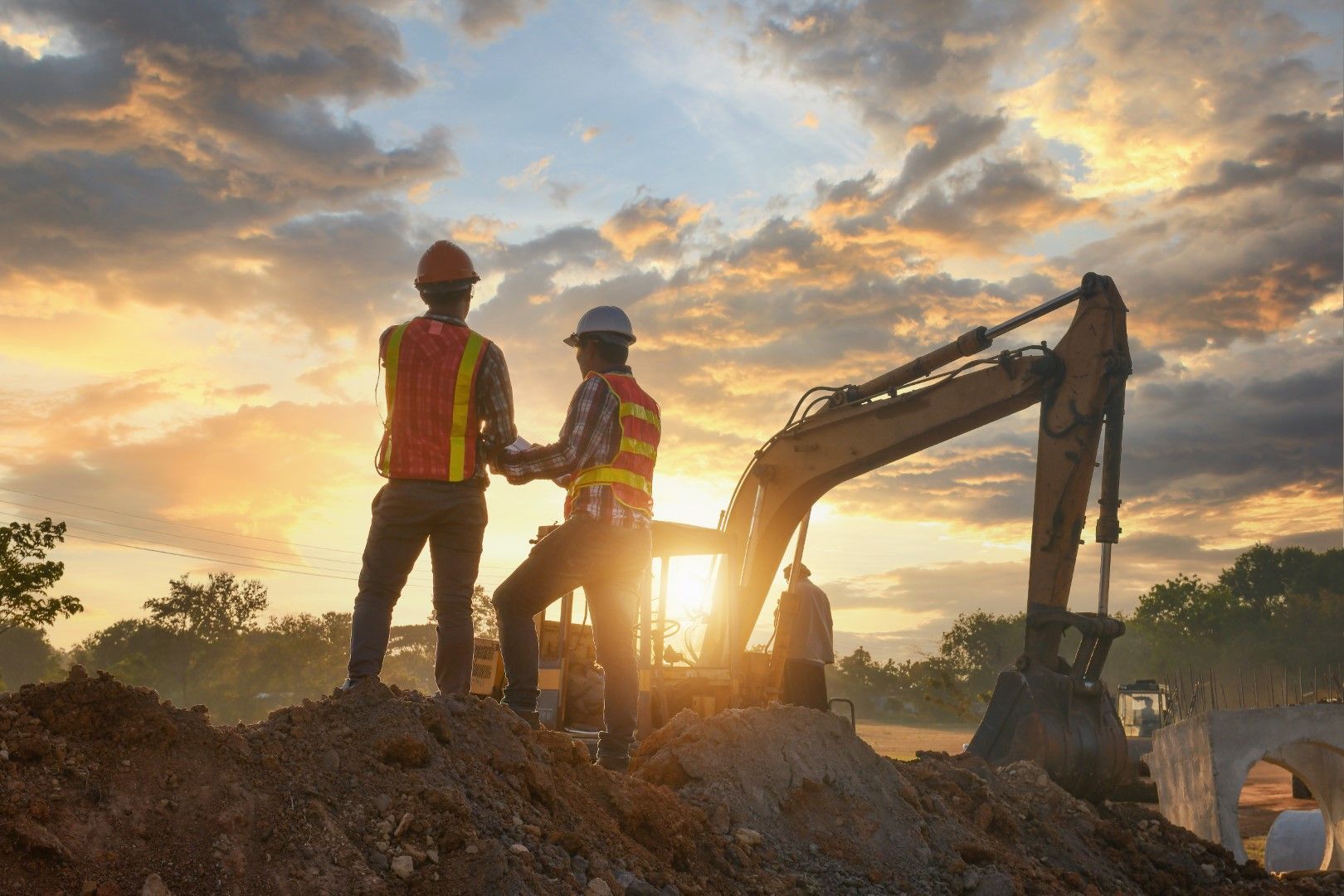 Two construction workers on a construction site looking at a plan at sunset next to an excavator.