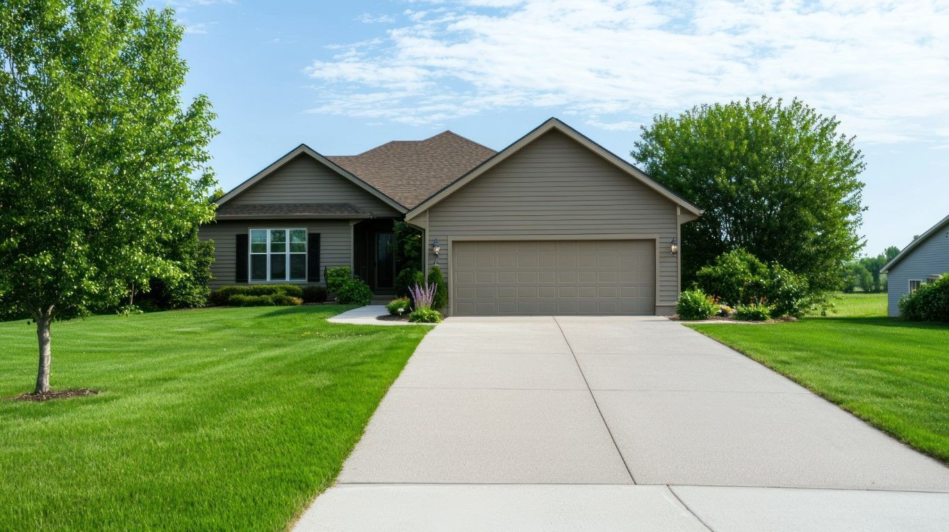 A one-story house with a brown exterior, a two-car garage, and a concrete driveway, set against a green lawn and a blue sky.