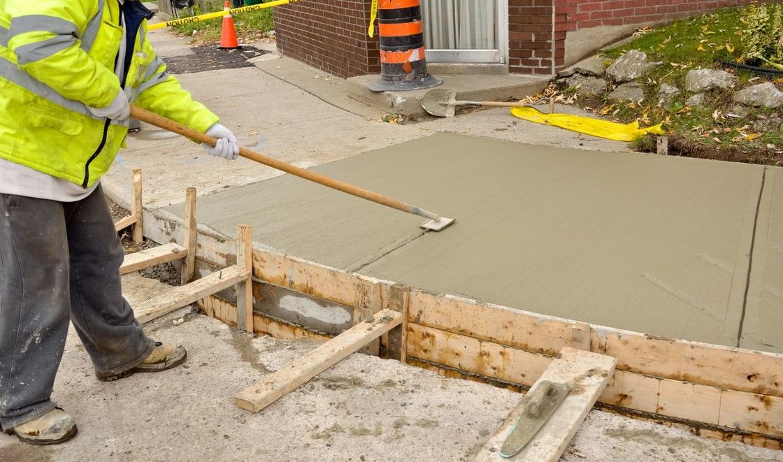 A construction worker levels fresh concrete with a long-handled tool in a wooden frame, wearing a yellow safety vest.
