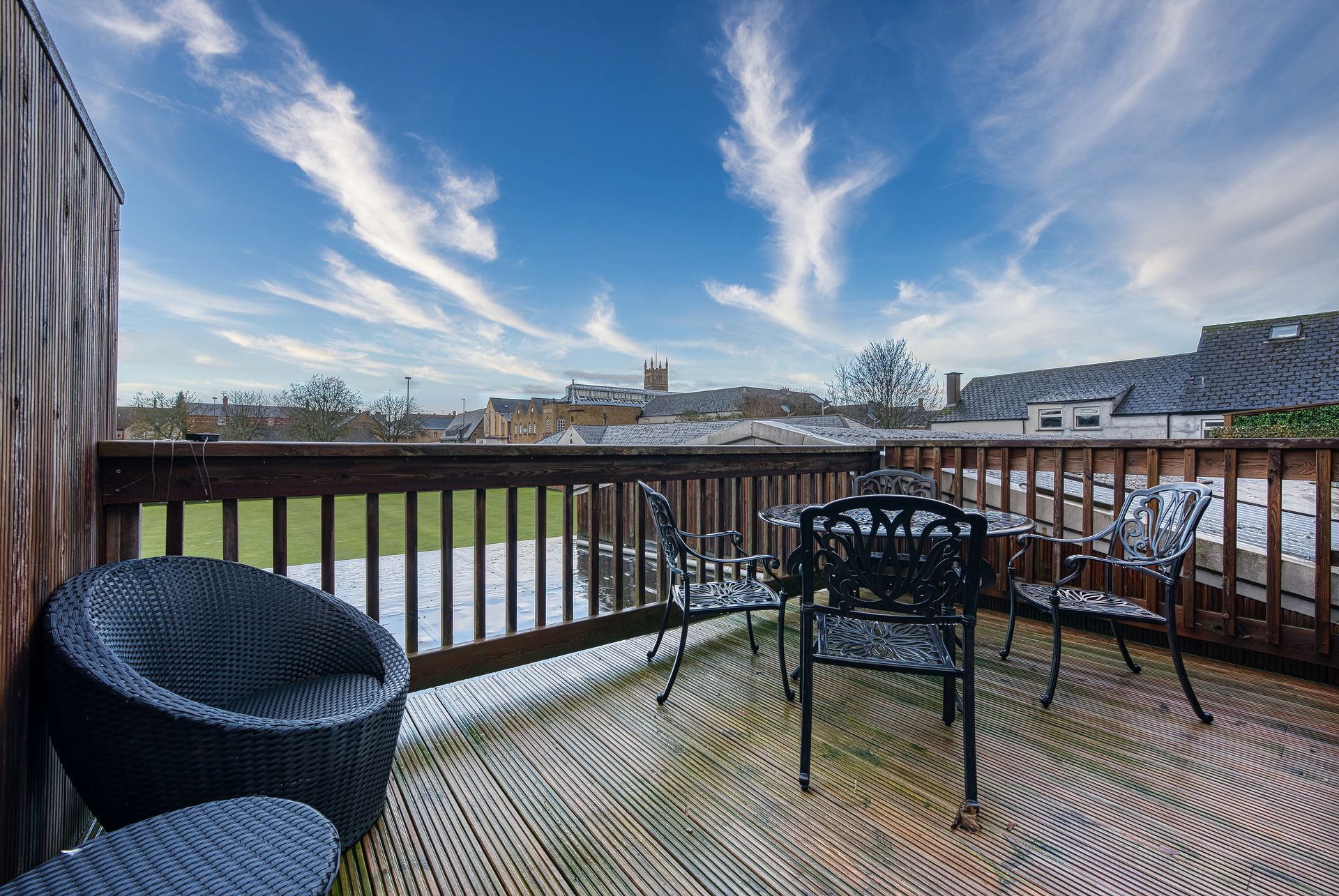 Wooden deck with outdoor furniture overlooking a city skyline under a blue sky with streaky clouds.