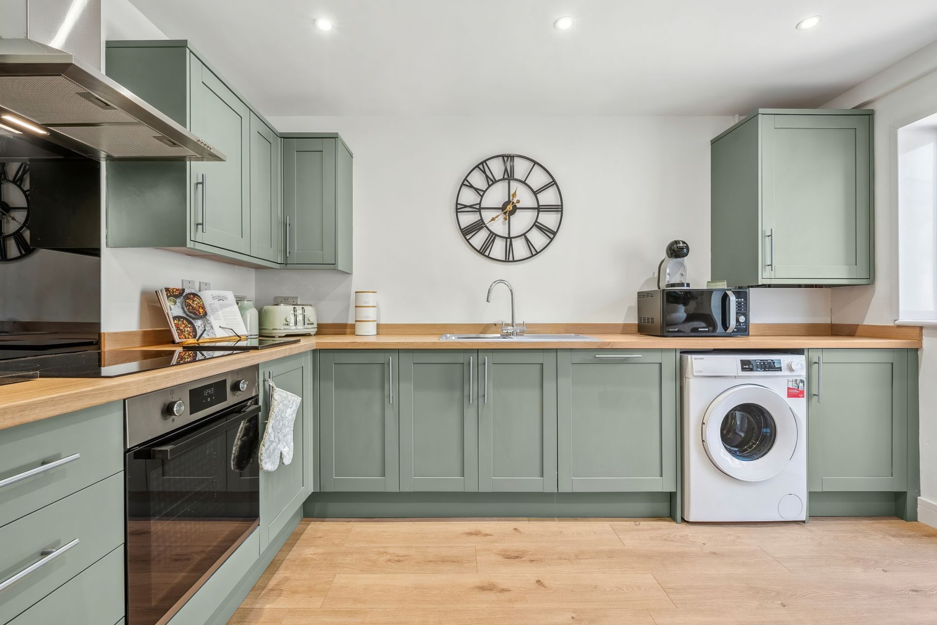 Green kitchen with wood countertops, stainless steel appliances, and a washing machine. A clock hangs on the wall.