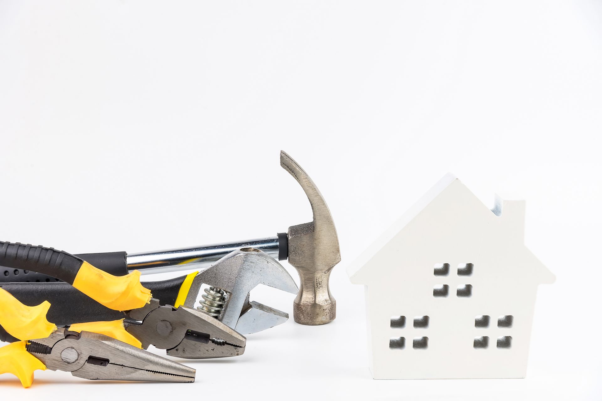 Tools including hammer, pliers, and wrench next to a white miniature house on a white surface.