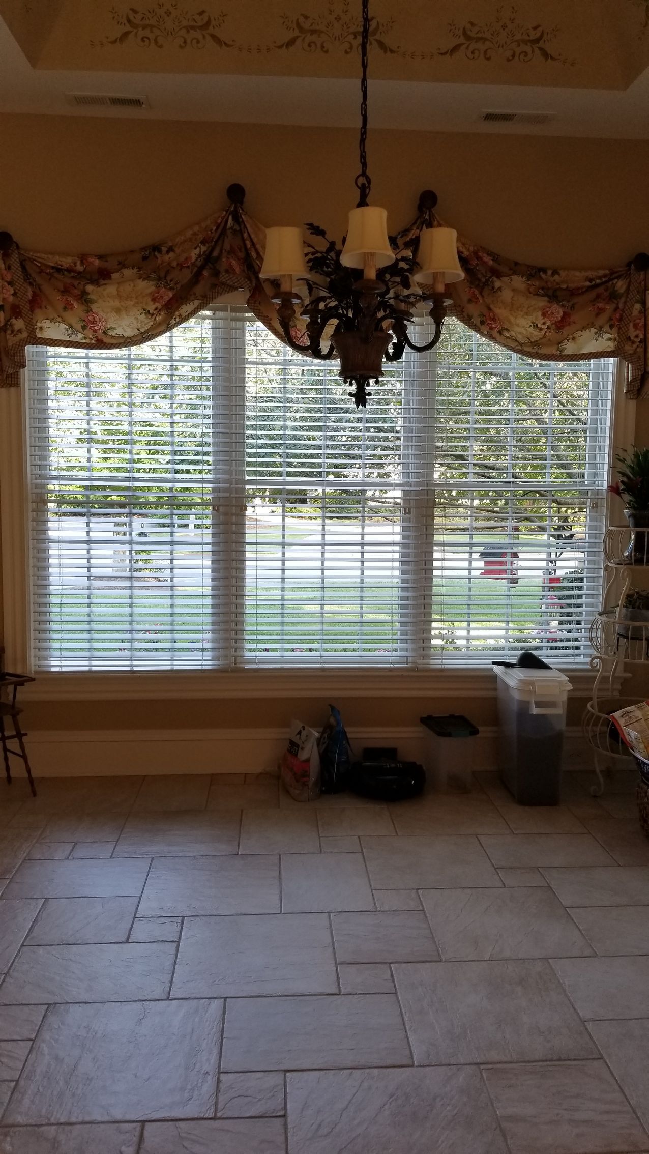 A room with large window, blinds, and ornate chandelier. Stone floor and decorative window valance.