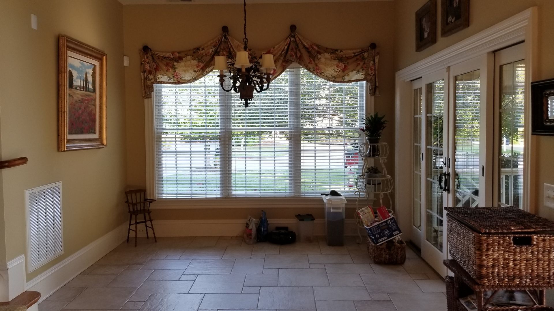 Entryway with window, chandelier, shutters, art, and basket. Beige walls and tiled floor.