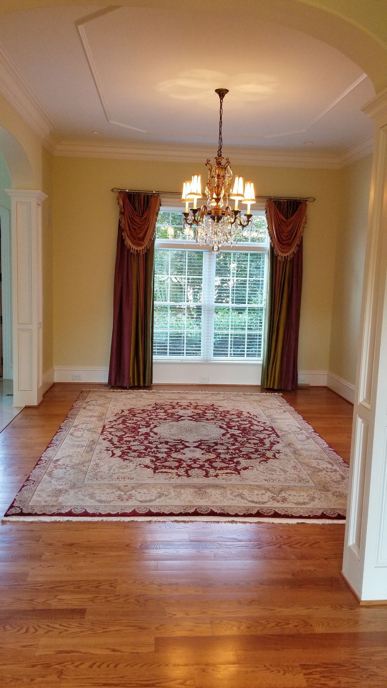 Dining room with chandelier, large rug, window with drapes, and wood flooring.