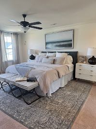 Bedroom with a large bed, bench, rug, artwork, and ceiling fan. Neutral tones with accents of black.
