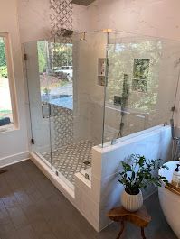 Glass shower enclosure in a modern bathroom, featuring white walls, gray floor, and a small wooden side table with a plant.