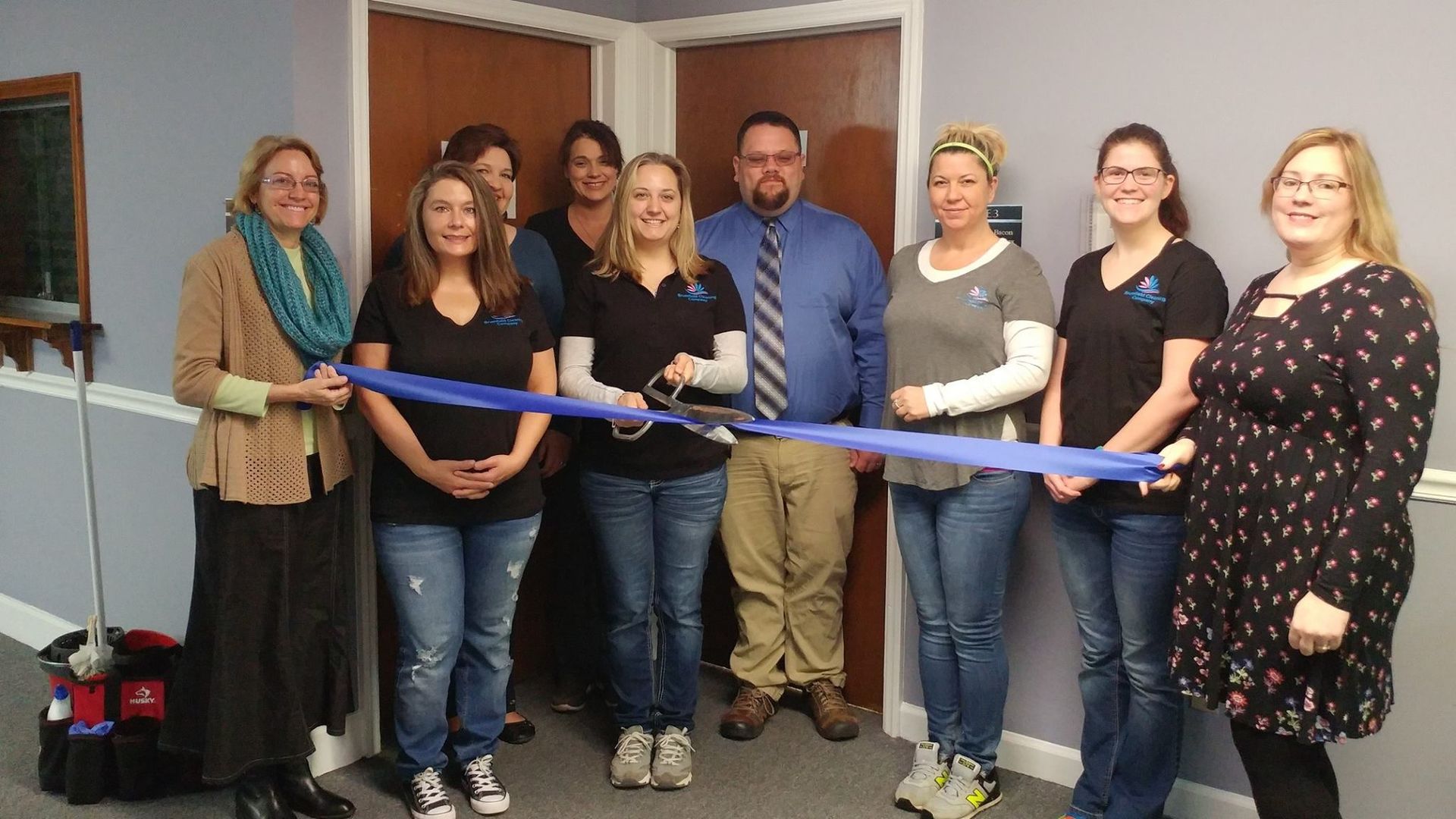 Group cuts blue ribbon at doorway; inside is visible. They are smiling, wearing casual clothes.