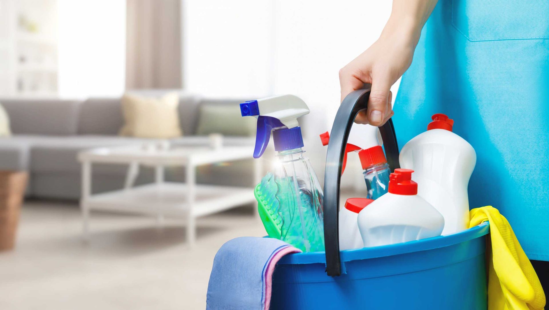 Hand holding a blue cleaning caddy with spray bottles and supplies in a living room.