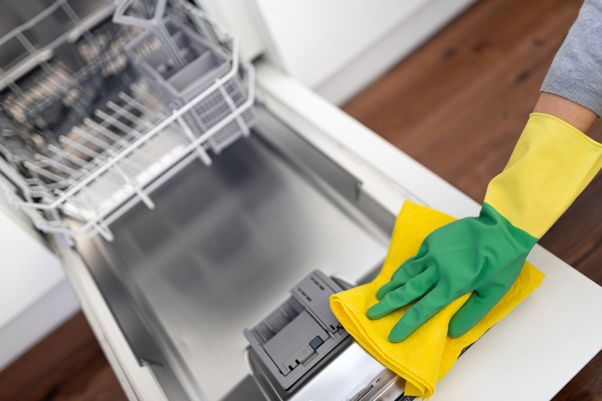 Person in yellow and green gloves cleaning the inside of an open dishwasher with a yellow cloth.