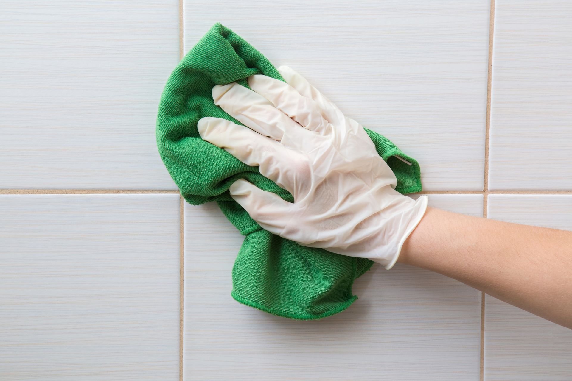 Gloved hand wipes a white-tiled wall with a green rag. Gloved hand wipes a white-tiled wall with a green rag.