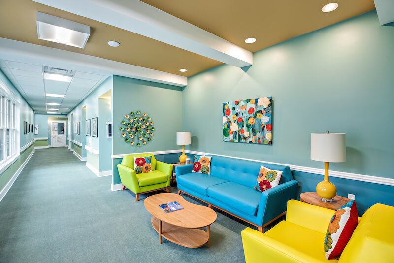 Lounge area with teal walls, aqua sofa, yellow and lime chairs, coffee table, and artwork; hallway in background.