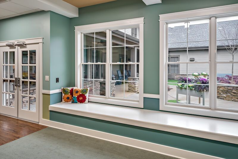 Interior view: teal walls, white-framed windows, window seat with pillow, French doors to the left, view of an outdoor area.