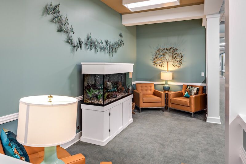 A waiting room with an aquarium, two orange chairs, and wall art against a blue-green wall.