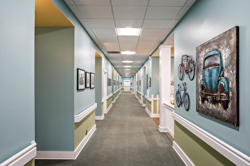 Long hallway with blue-green walls, artwork, and doorways, typical of a care facility.