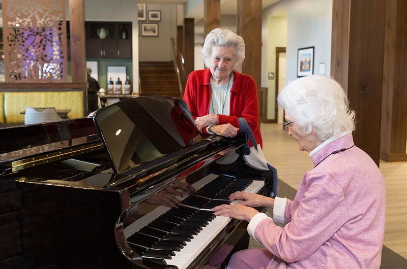 Two elderly women at a piano; one plays, the other watches. Interior setting, well-lit, wood accents.