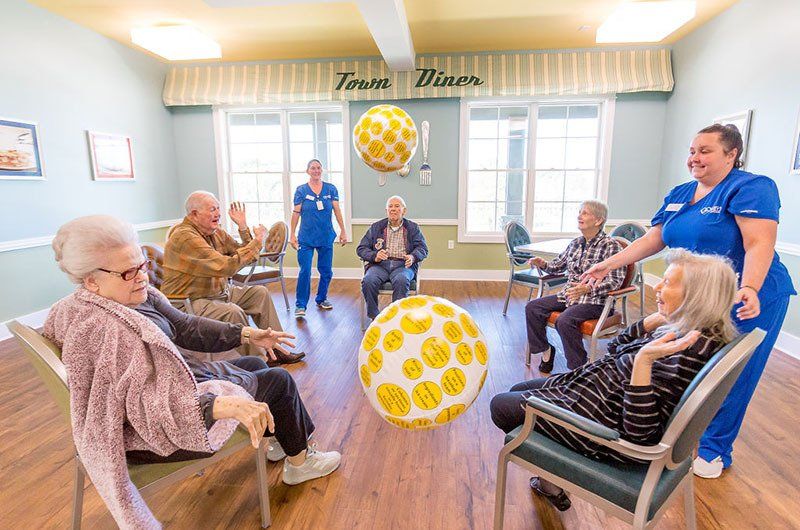 Seniors playing with giant yellow smiley face balls in a room, with two nurses present.