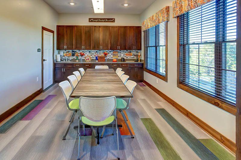 A long wooden table in a brightly lit room, with cabinets, windows and chairs.