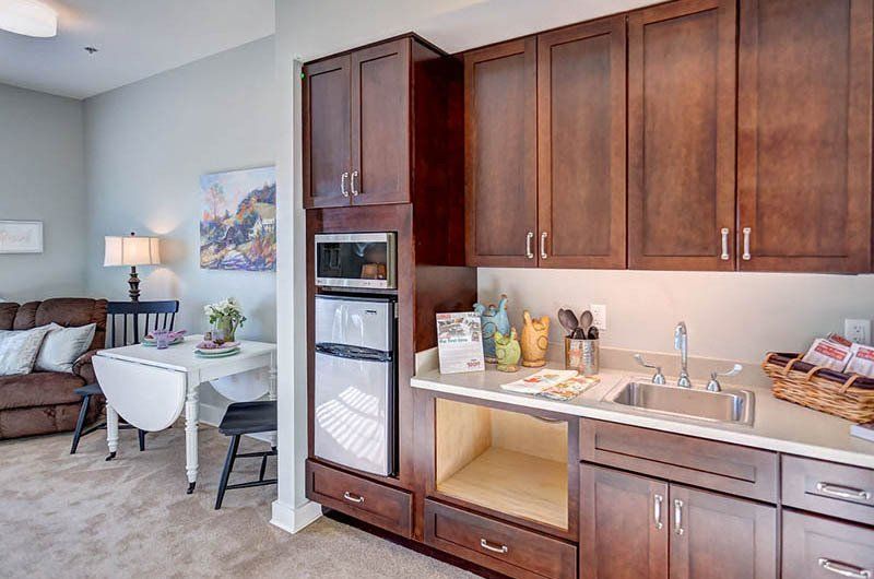 Kitchenette with dark cabinets, microwave, sink, and small refrigerator. Table and sofa in background.