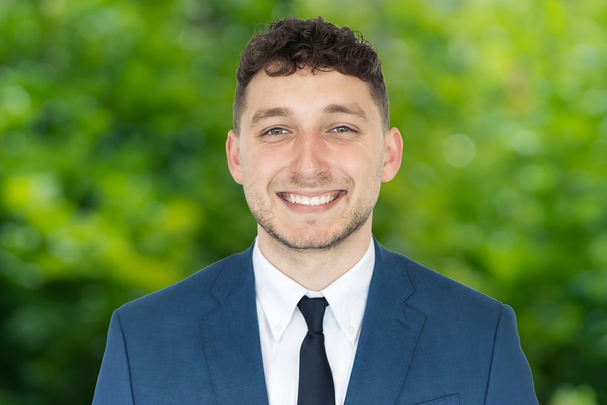Man in blue suit smiling, set against a blurred green backdrop of foliage.