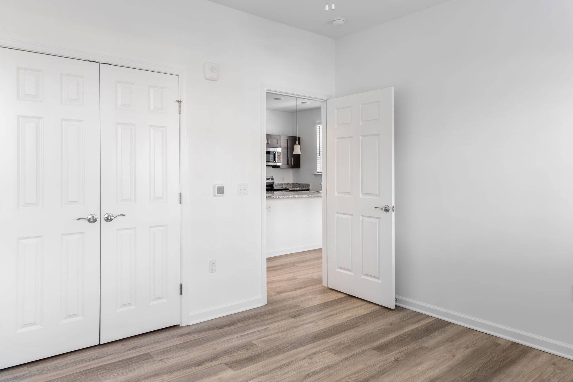 Empty bedroom with white doors, closet, and entryway to a kitchen with cabinets and microwave.