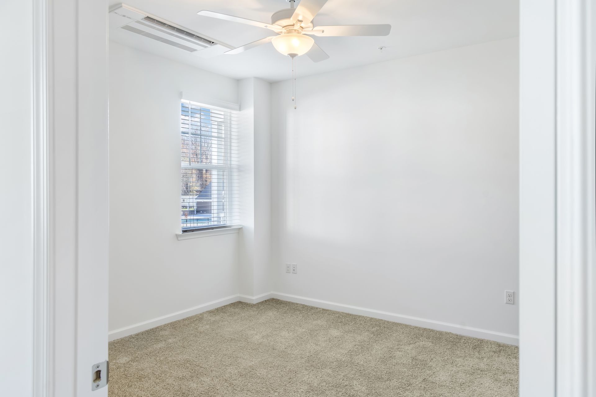 Empty, white-walled room with a beige carpet, window with blinds, and ceiling fan.