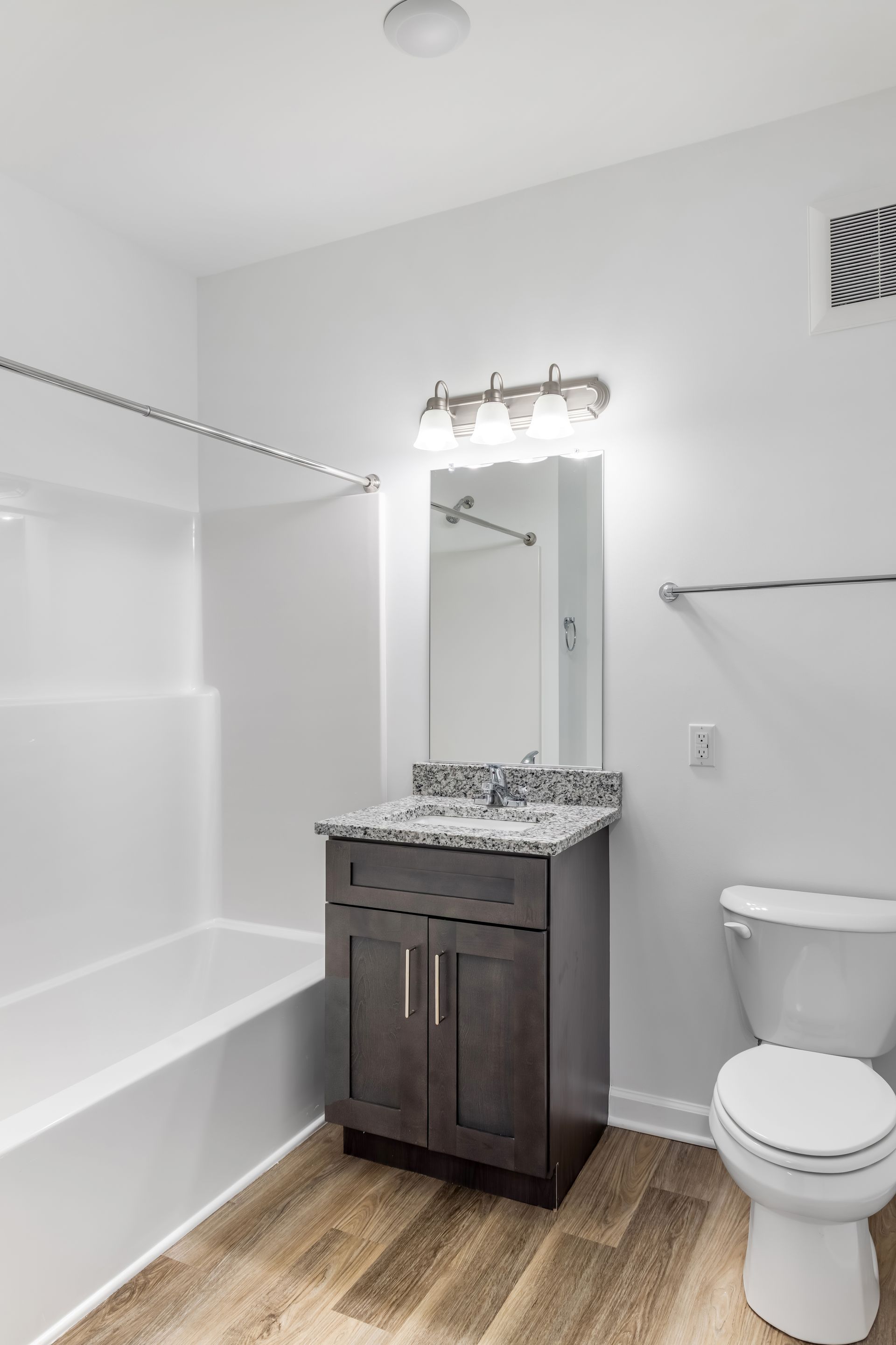 Bathroom with white tub, toilet, vanity, and a mirror.  Brown wooden floor and white walls.