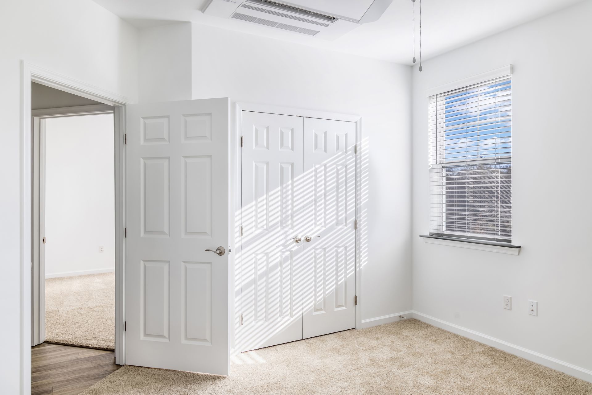 Empty white bedroom with closed closet, open door, and window with blinds.